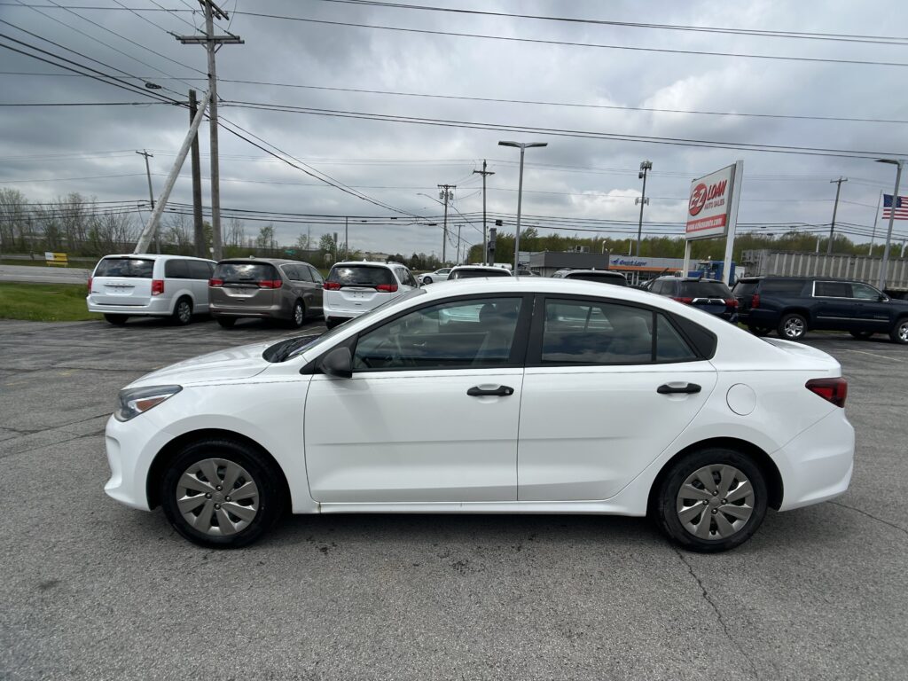 White sedan in the foreground of a car lot with several minivans and a large dealership sign under an overcast sky