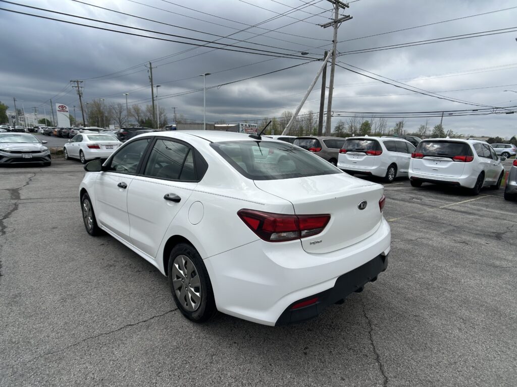 White Kia Rio in the foreground at a crowded car dealership lot, with many white cars and dark, cloudy skies above.
