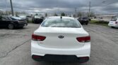 Rear view of a white Kia Rio in a parking lot with other vehicles and a cloudy sky above.