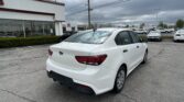 White Kia sedan in a dealership lot with a white building and many cars in the background under a cloudy sky.