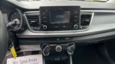Inside a car cabin showing the center dashboard with a touchscreen displaying 12:04 PM, surrounded by control buttons; outside the windshield is a busy car dealership lot with many parked cars and signage.