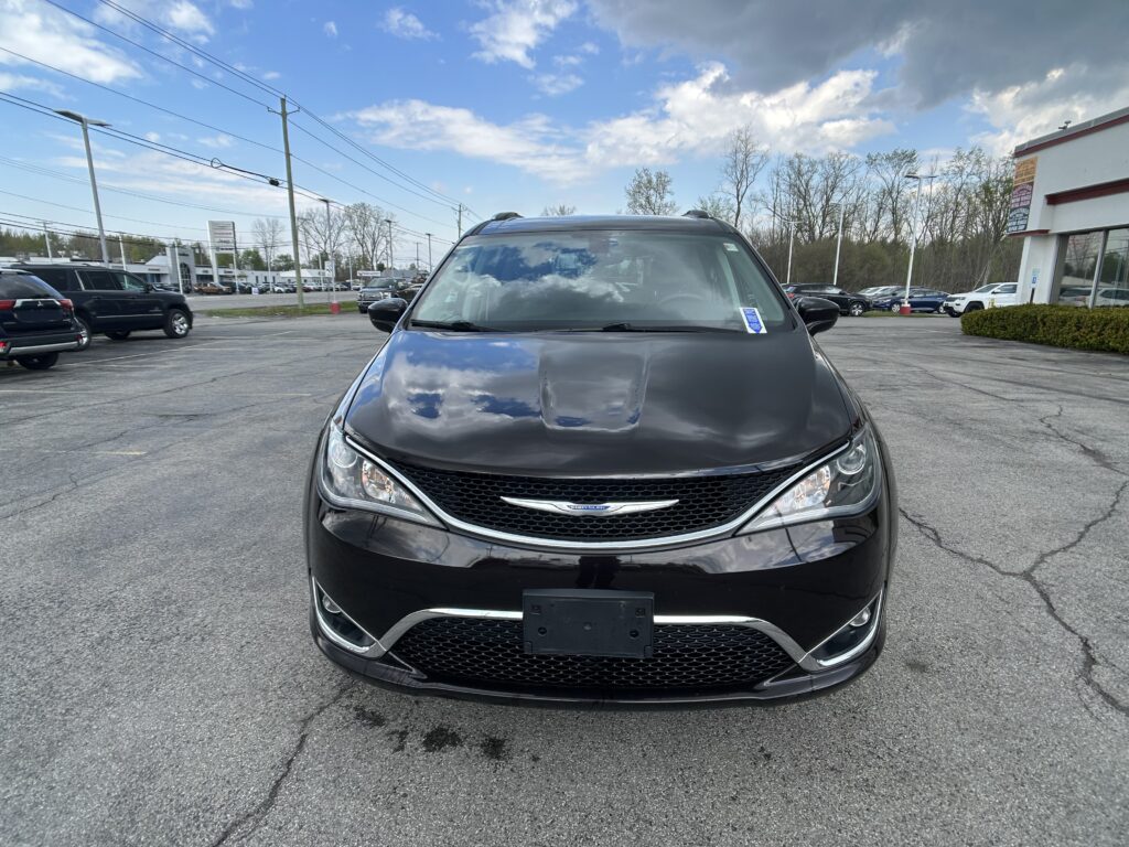 Front view of a black Chrysler minivan parked in a dealership lot, with other cars and a building on the right.