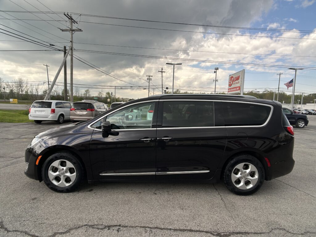 Black minivan in a dealership lot with other cars, power lines overhead, and a cloudy sky in the background.