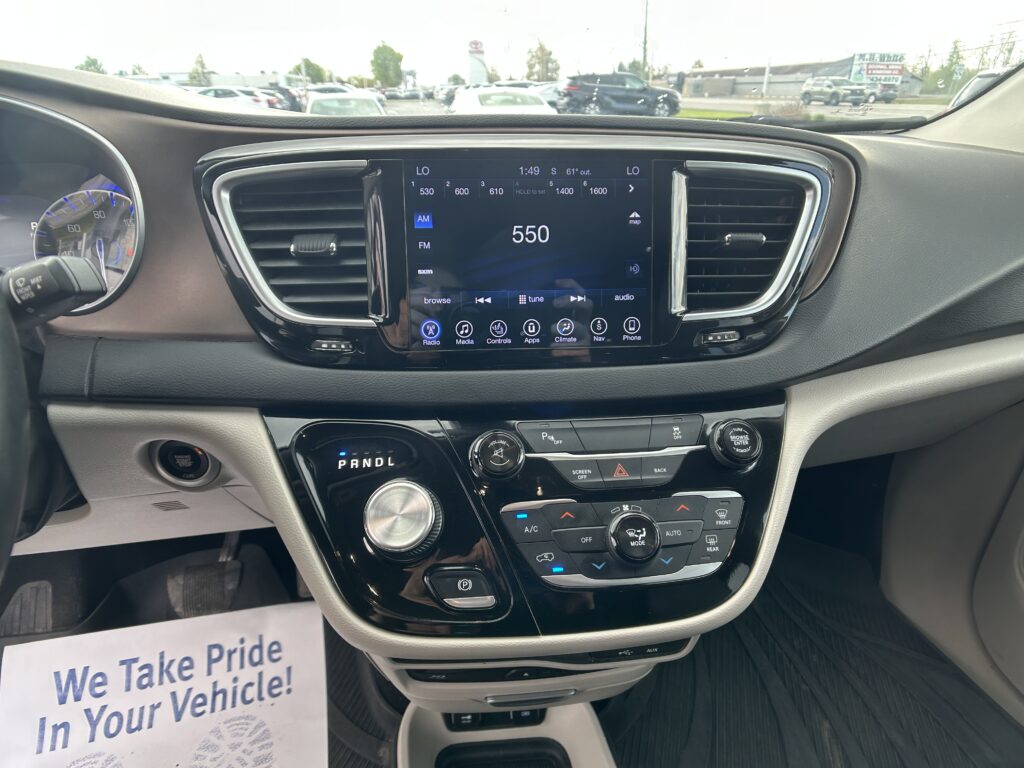 Center console infotainment screen displaying 550 AM radio with climate and control icons, surrounded by the car’s dashboard and vents. Outside through the windshield is a busy dealership lot.
