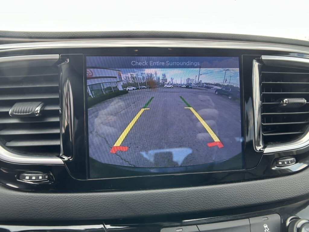 In-dash rearview camera display showing parking guide lines and the street behind the car; text 'Check Entire Surroundings' appears on screen.