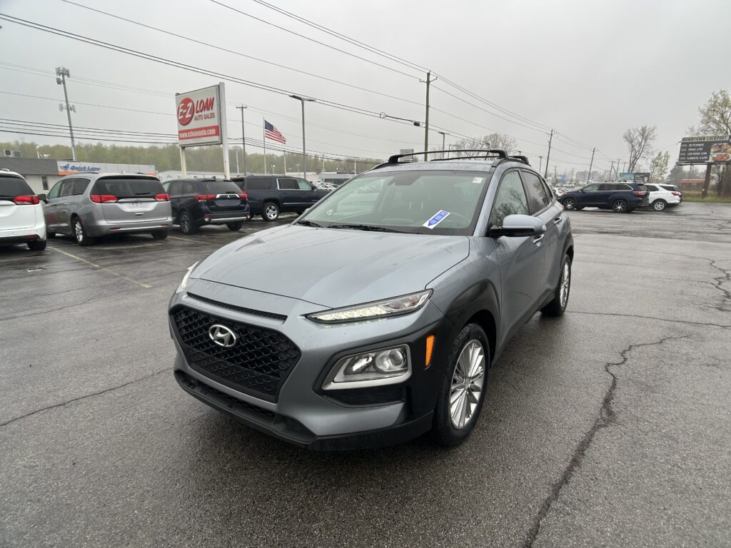 Front-right view of a gray Hyundai SUV in a rainy parking lot with other cars and an E-Z Loan Auto Sales sign in the distance. American flag nearby.