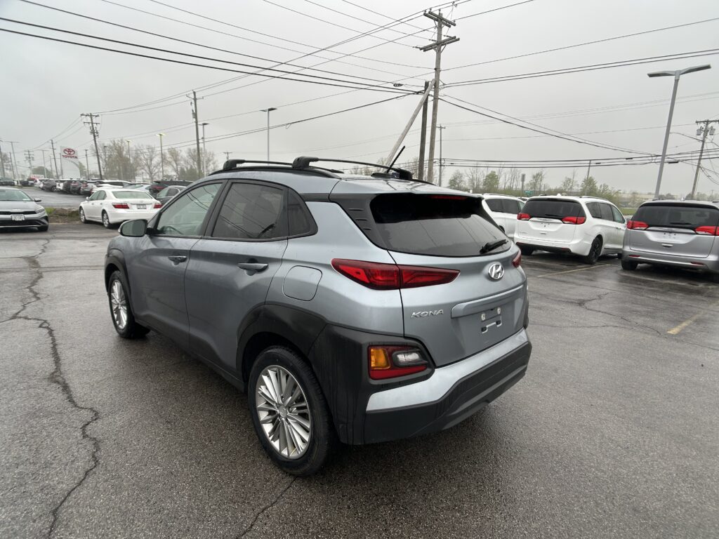 Rear three-quarter view of a silver Hyundai Kona in a crowded dealership lot with other cars nearby and power lines overhead.