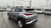 Rear three-quarter view of a silver Hyundai Kona in a crowded dealership lot with other cars nearby and power lines overhead.