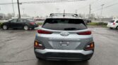 Rear view of a silver Hyundai Kona SUV in a parking lot with other cars and overhanging power lines above.