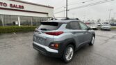 Rear three-quarter view of a gray Hyundai SUV parked in a dealership lot with a white building and red trim in the background.