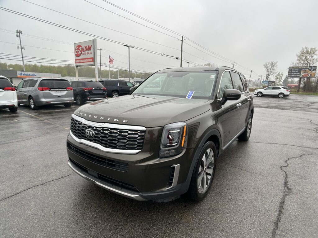 Front-right three-quarter view of a gray Kia Telluride parked in a busy dealership lot, with a large EZ LOAN sign and power lines in the background.