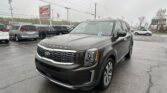 Front-right three-quarter view of a gray Kia Telluride parked in a busy dealership lot, with a large EZ LOAN sign and power lines in the background.