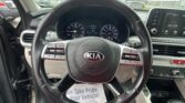 Driver's view of a Kia interior: steering wheel centered, instrument cluster, and dashboard touchscreen to the right; outside shows a parking lot through the windshield.