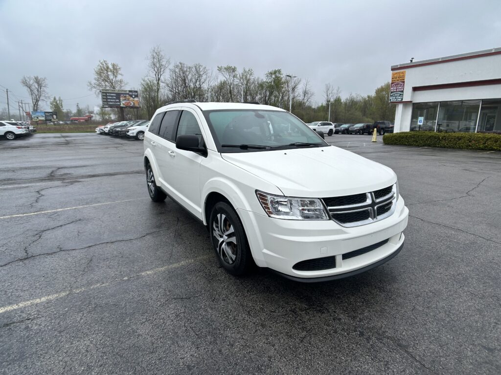 White SUV in the foreground of a wet parking lot, with a dealership building on the right and other cars in the background on a cloudy day.