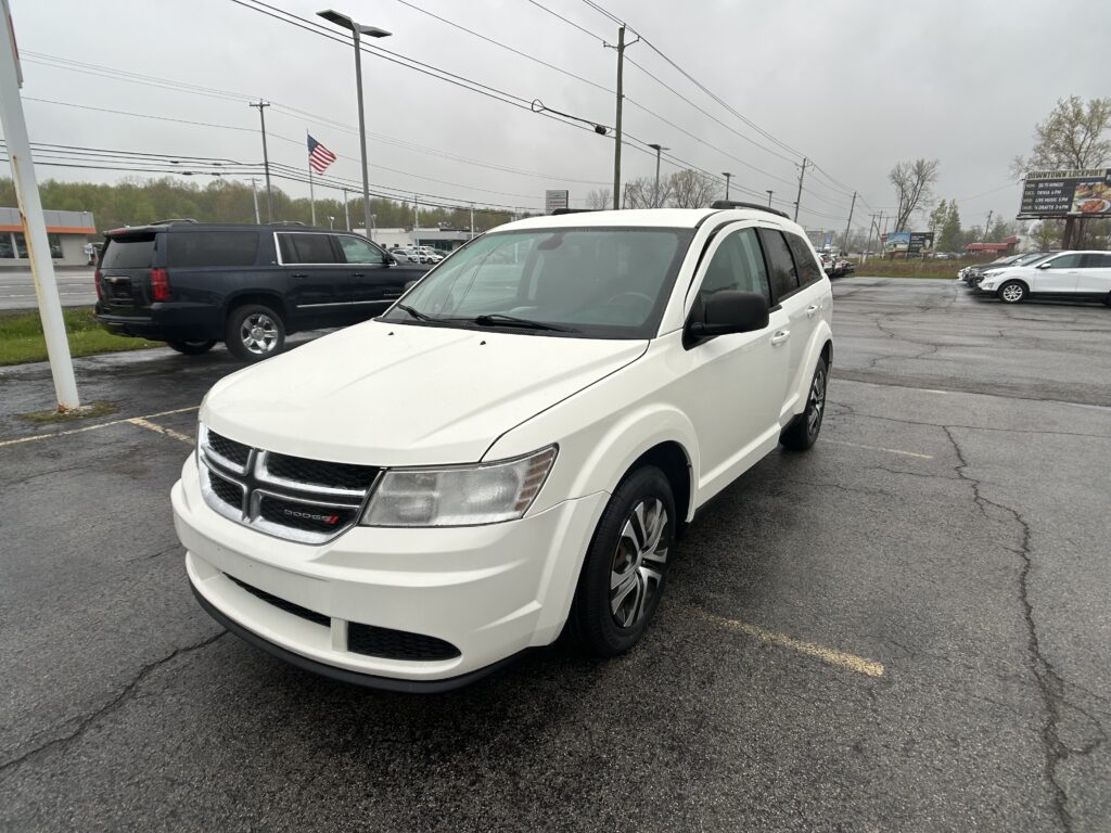 White Dodge SUV in a dealership lot, front angled view with other vehicles and an American flag in the background on a cloudy day.