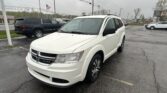 White Dodge SUV in a dealership lot, front angled view with other vehicles and an American flag in the background on a cloudy day.