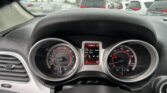 View from the driver seat of a car dashboard with tachometer, speedometer, and a red digital display, while a rainy parking lot is visible through the windshield.