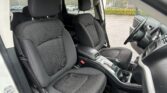 Interior of a vehicle: driver and front passenger seats with black patterned upholstery, center console, and steering wheel; parking lot visible outside the windows.
