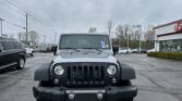 Front view of a silver Jeep Wrangler in a parking lot, dealership visible on the right under an overcast sky.