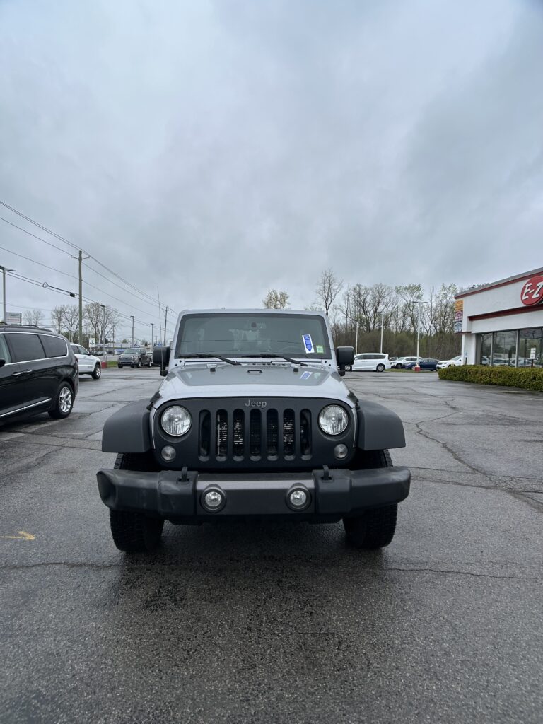 Front view of a silver Jeep Wrangler in a parking lot, dealership visible on the right under an overcast sky.