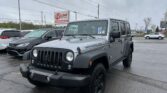 Front view of a gray Jeep Wrangler parked in a car dealership lot, with other cars and an EZ Loan sign in the background.