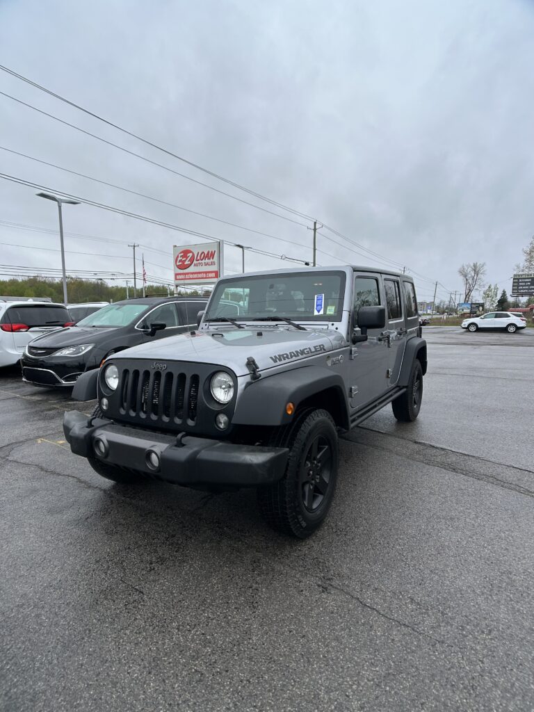 Front view of a gray Jeep Wrangler parked in a car dealership lot, with other cars and an EZ Loan sign in the background.
