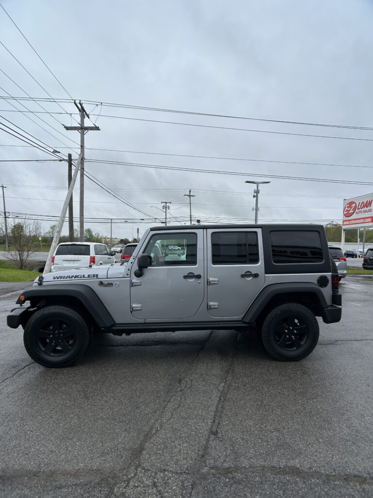 Silver Jeep Wrangler SUV parked in a lot with utility poles and overhead power lines, and an EZ LOAN Auto Sales sign in the background.