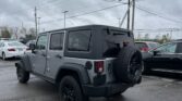 Gray Jeep Wrangler with a rear-mounted spare tire parked in a lot under a cloudy sky and utility lines overhead