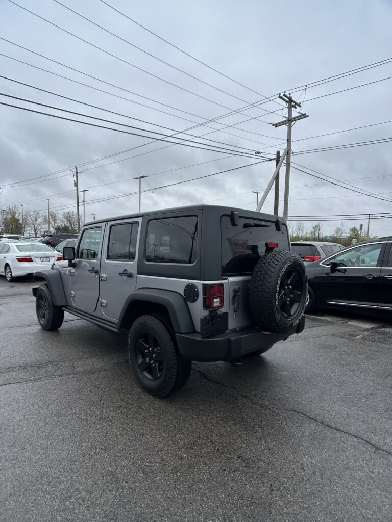 Gray Jeep Wrangler with a rear-mounted spare tire parked in a lot under a cloudy sky and utility lines overhead