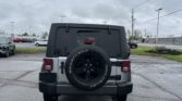 Rear view of a gray Jeep with spare tire mounted on the back in a parking lot, overcast sky and power lines above.