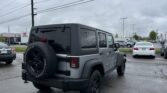 Gray Jeep Wrangler with spare tire mounted on rear, parked in a car dealership lot on a cloudy day.
