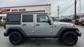 Silver Jeep Wrangler parked in front of a white auto dealership building on a cloudy day in a car lot.