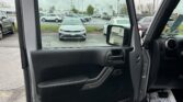 Interior view of an open car door with a black panel; a dealership parking lot with cars is visible through the window frame in the background.