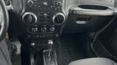 View of a black Jeep interior: center console with radio, climate controls, gear shifter, cupholders, and front passenger seat; parking lot visible through windshield.