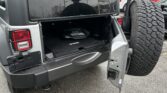 Rear of a gray Jeep Wrangler with the tailgate open and a mounted spare tire visible on the back, showing the V6 engine badge in the cargo area.