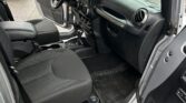 Interior of a Jeep Wrangler: driver’s seat, steering wheel, center console, and dashboard seen through an open door, dealership lot outside.