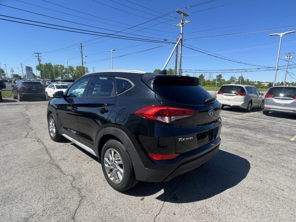 Rear three-quarter view of a black Hyundai Tucson SUV in a sunny dealership lot with other cars and overhead power lines in the background.
