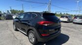 Rear three-quarter view of a black Hyundai Tucson SUV in a sunny dealership lot with other cars and overhead power lines in the background.