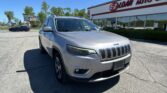 Silver Jeep SUV in the foreground at an auto dealership lot with a white building and red signage in the background under a clear blue sky.