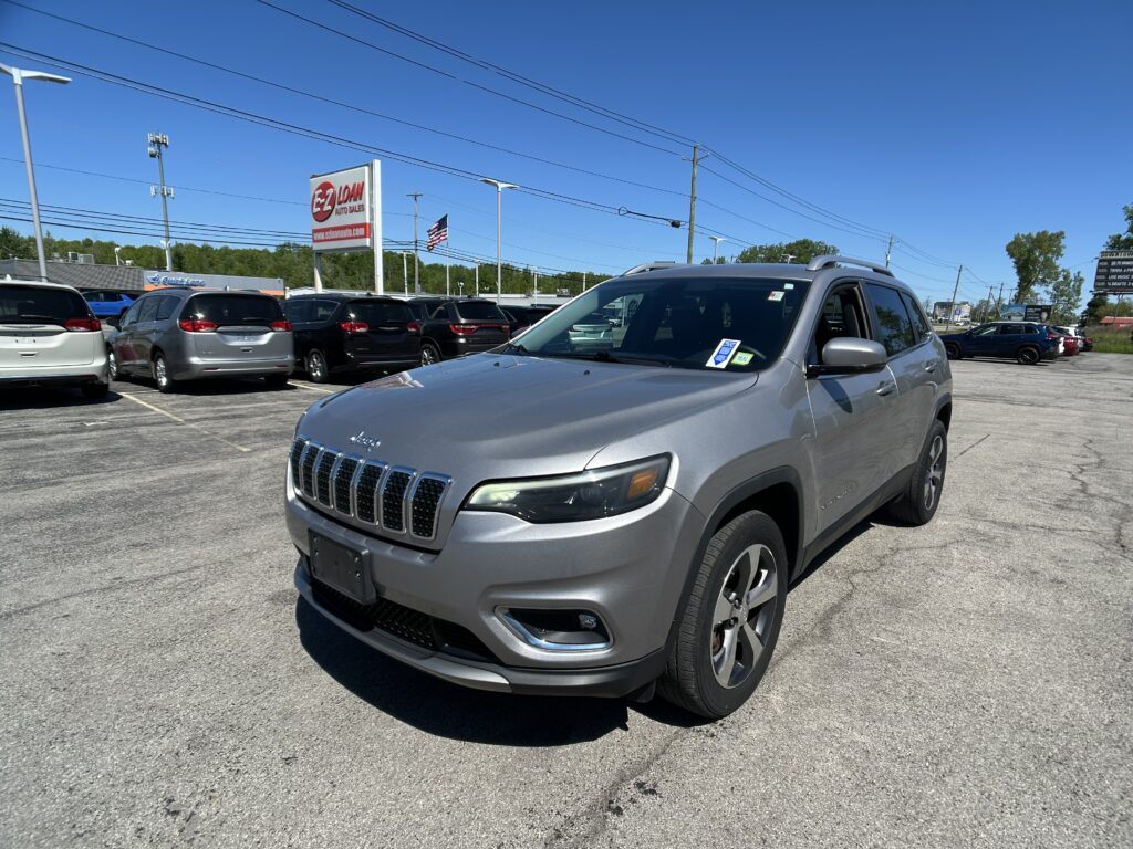 Front-right view of a silver Jeep SUV in a car dealership lot with many parked cars and a red EZ Loan Auto Sales sign in the distance, under a clear blue sky.