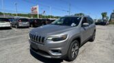 Front-right view of a silver Jeep SUV in a car dealership lot with many parked cars and a red EZ Loan Auto Sales sign in the distance, under a clear blue sky.