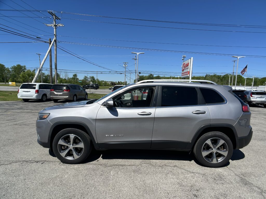 Silver SUV in the foreground at a car dealership lot with several other vehicles, a tall utility pole and power lines overhead against a clear blue sky, and an American flag in the distance.