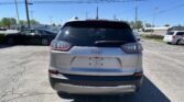 Rear view of a silver Jeep SUV with '4x4' and 'Limited' badges parked in a lot, other cars and power lines in the background under a blue sky.