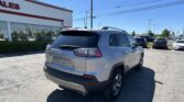 Rear view of a silver Jeep Grand Cherokee in a dealership parking lot with a white building and 'SALES' sign, under a clear blue sky.