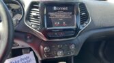 Interior of a car showing a center touchscreen (Uconnect) with climate and driving controls, seen from the driver's seat, on a sunny day; papers on the floor read 'We Take Pride In Your Vehicle.'