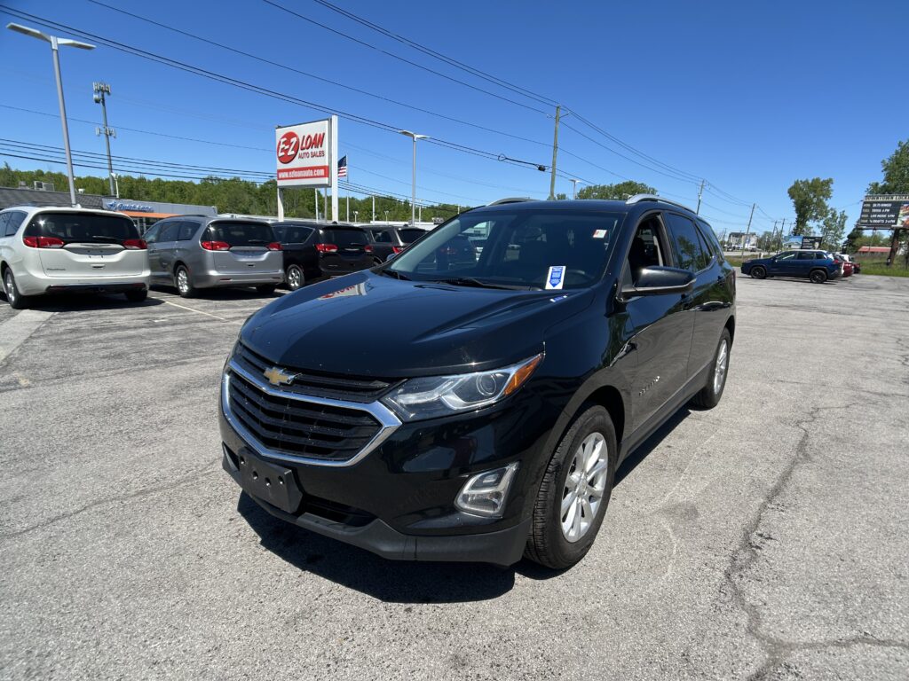 Black Chevrolet SUV in foreground at a car lot with other parked minivans and a tall EZ LOAN Auto Sales sign in the background on a sunny day