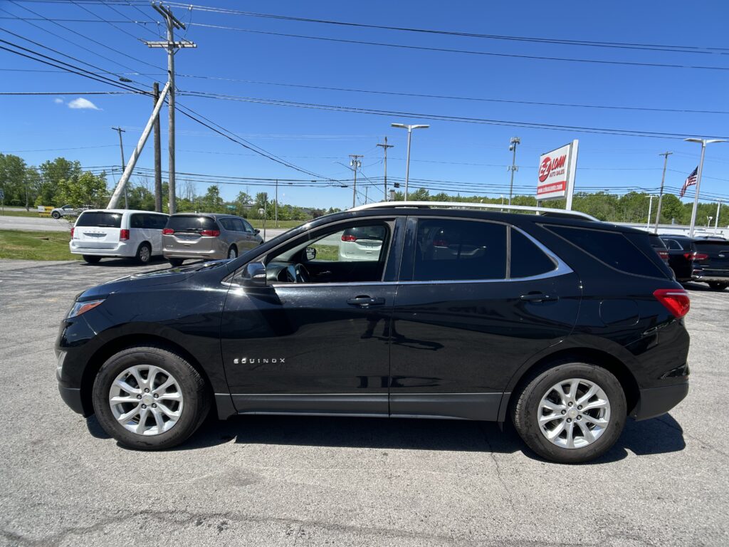 Black Chevrolet Equinox parked in a dealership lot with other vehicles, power lines overhead and a sign in the background on a sunny day.