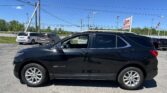 Black Chevrolet Equinox parked in a dealership lot with other vehicles, power lines overhead and a sign in the background on a sunny day.