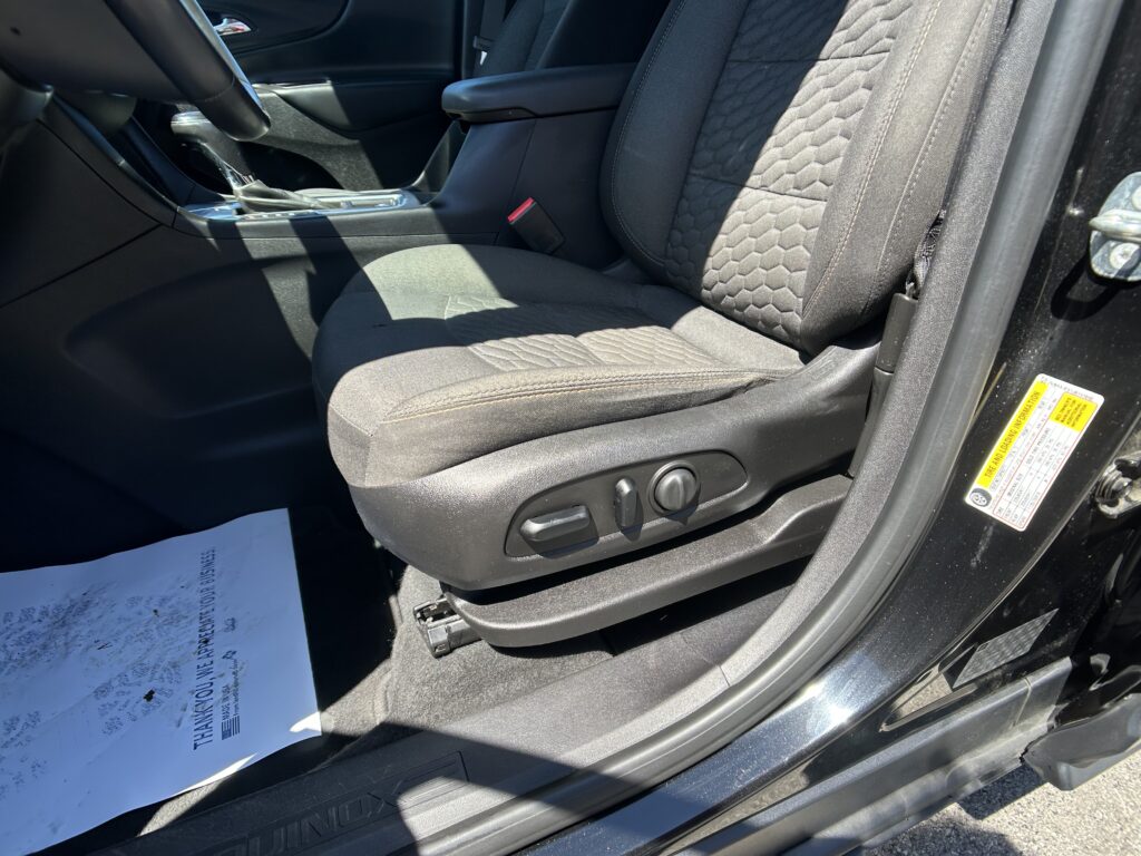 Front interior of a car showing the driver seat and center console, with a red seat belt buckle and textured seat fabric in dark tones.
