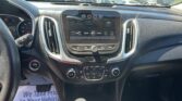 Car dashboard view with a central touchscreen display showing radio info, climate controls below, and a gear shift in the foreground; outside parking lot visible through windows.